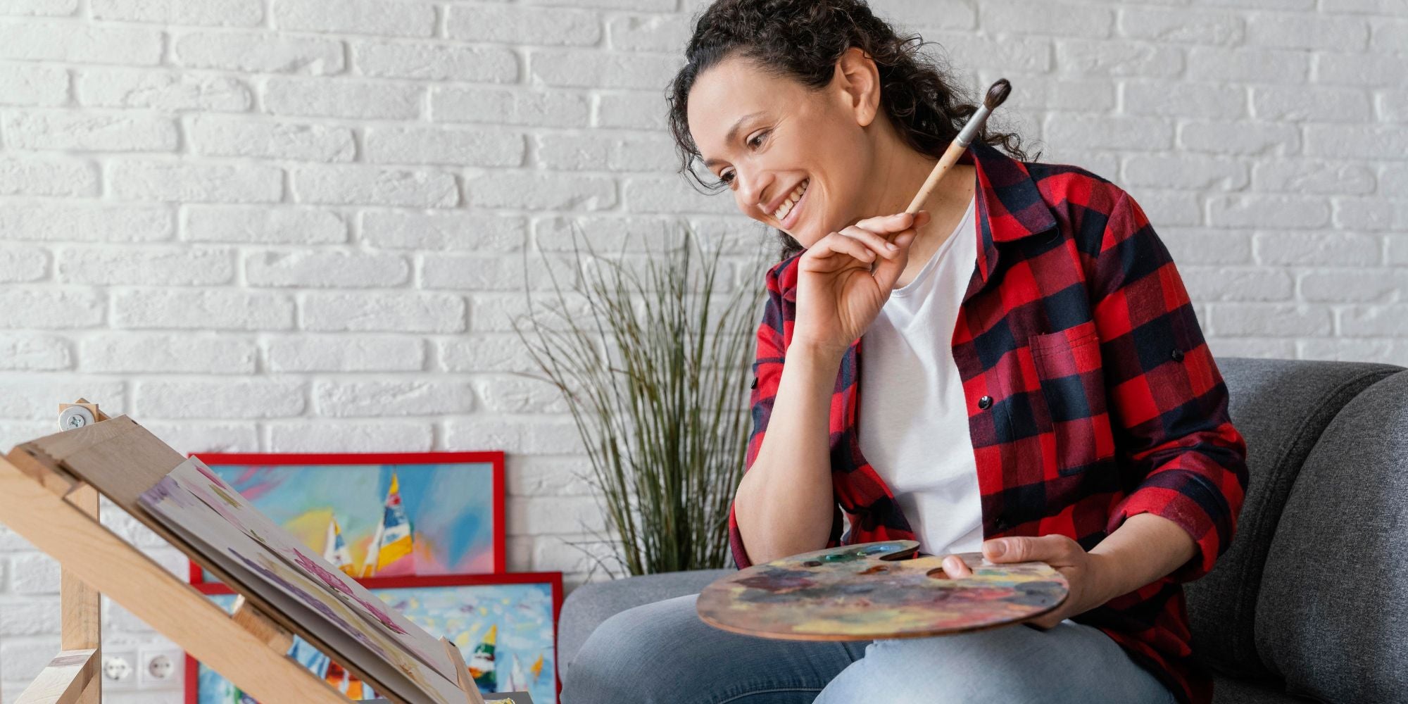 Woman sitting on a couch holding a paintbrush and palette, with art supplies and paintings in the background.